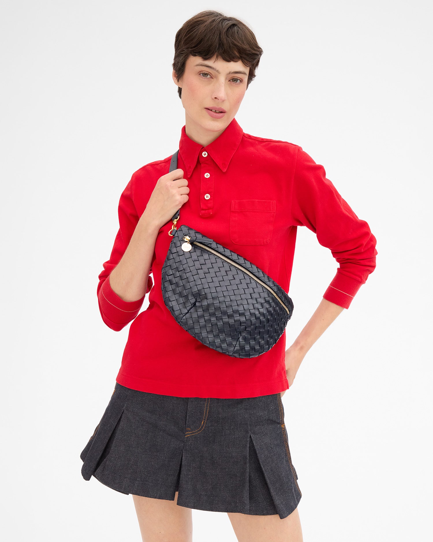 Model wearing a red long-sleeve top with  a denim pleated skirt and carrying the Twilight Diagonal Woven Grande Fanny over and across her chest. 